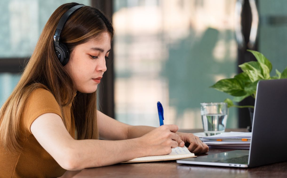 young female student wearing headphones taking notes on a notepad while sitting at a desk with a laptop