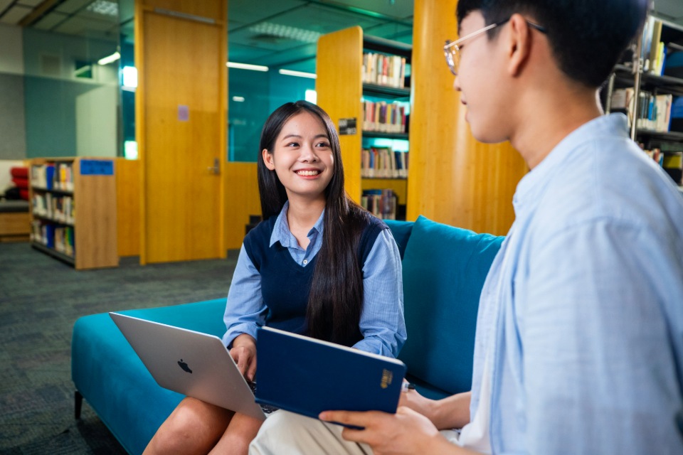 Two students sitting on a couch in a library, smiling and discussing while using a laptop and tablet
