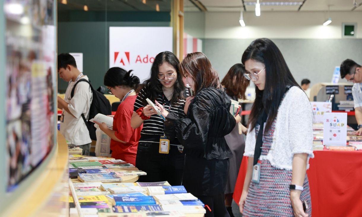  Students browse and read books displayed on tables during a campus book swap event, with an Adobe sign in the background.