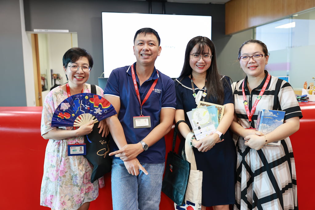 Four people with RMIT lanyards pose indoors in front of a red counter; two hold books and one holds a decorative fan. Their faces are smiling. 