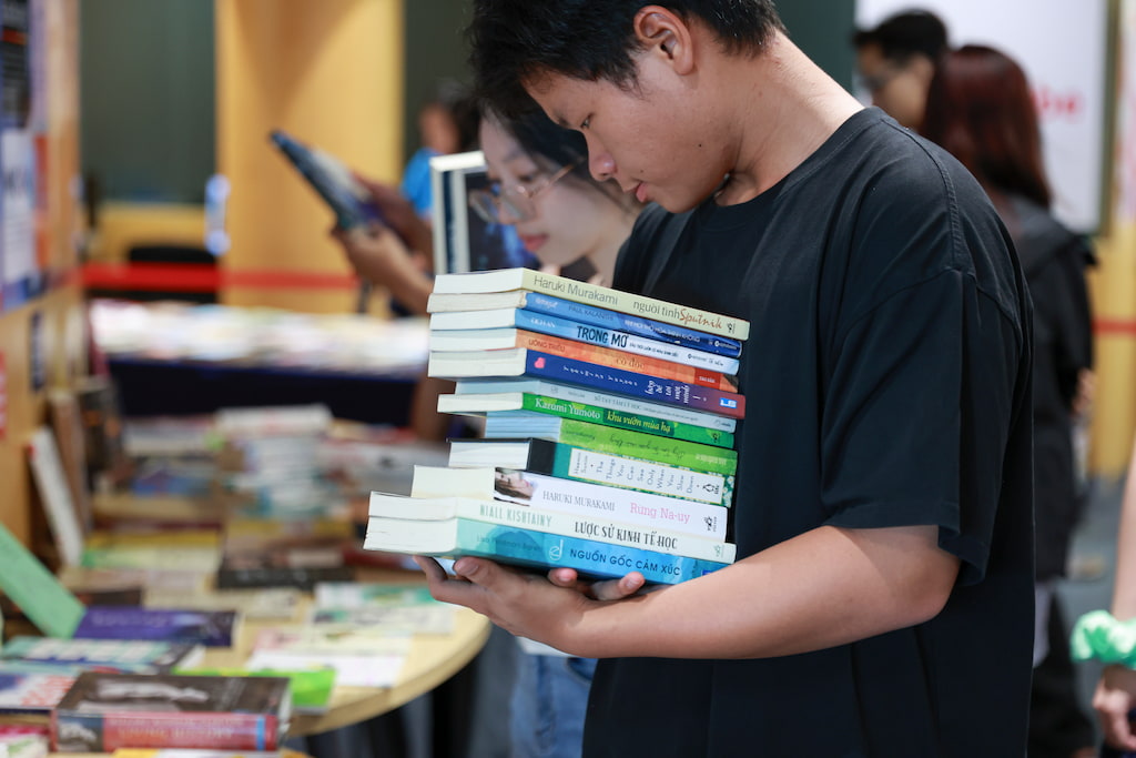 A person in a black shirt holds a large stack of books in their arms at an indoor event, with more books displayed on tables nearby. 