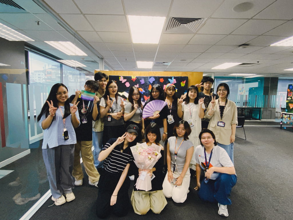A group of students with RMIT lanyards pose indoors in front of a decorated board, some holding fans and flowers, making peace signs
