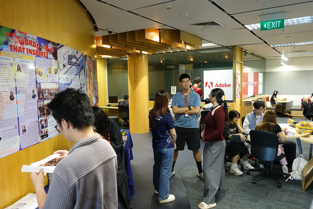 Students interact and read posters in a modern campus space, with some seated and some standing near an "Adobe" branded area.