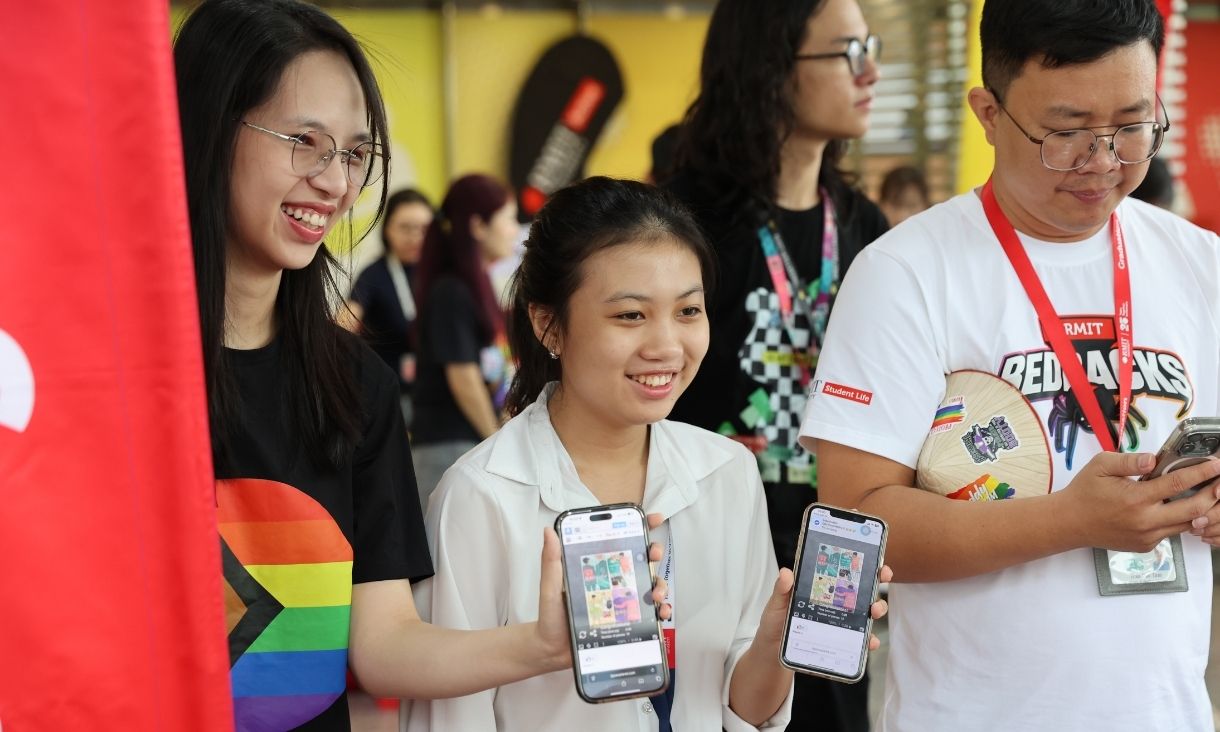 The photo of a group of 3 people, smiling while showing their phones out. 