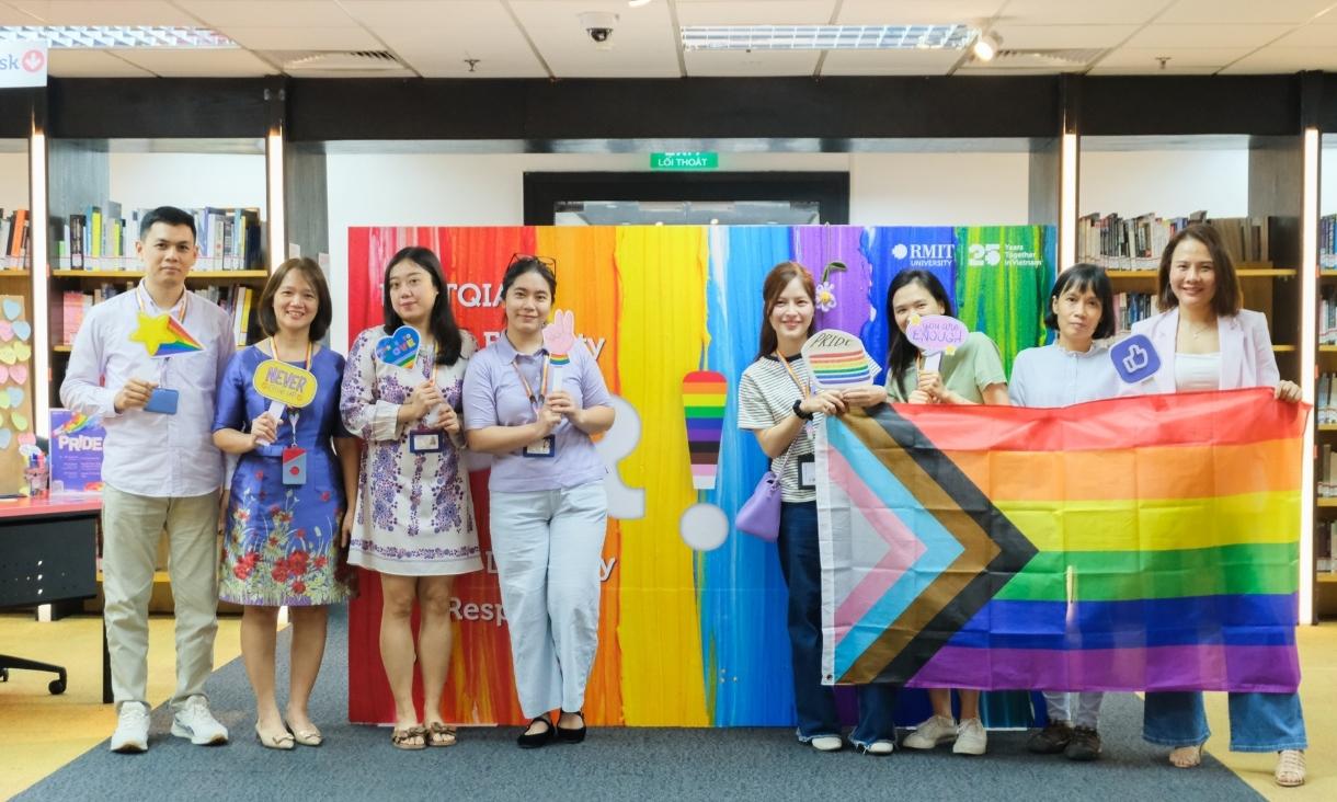 The photo of 8 people, all smile at the camera, taking a group shot photo with the PRIDE flag and props.