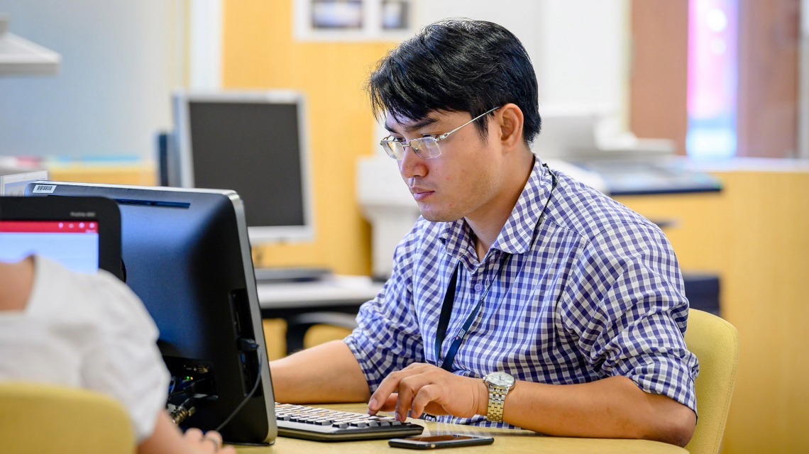 Image of Thi Nguyen, short dark hair, silver thin framed rectangular glasses and a navy blue tshirt in three quarter pose facing the camera and smiling. 