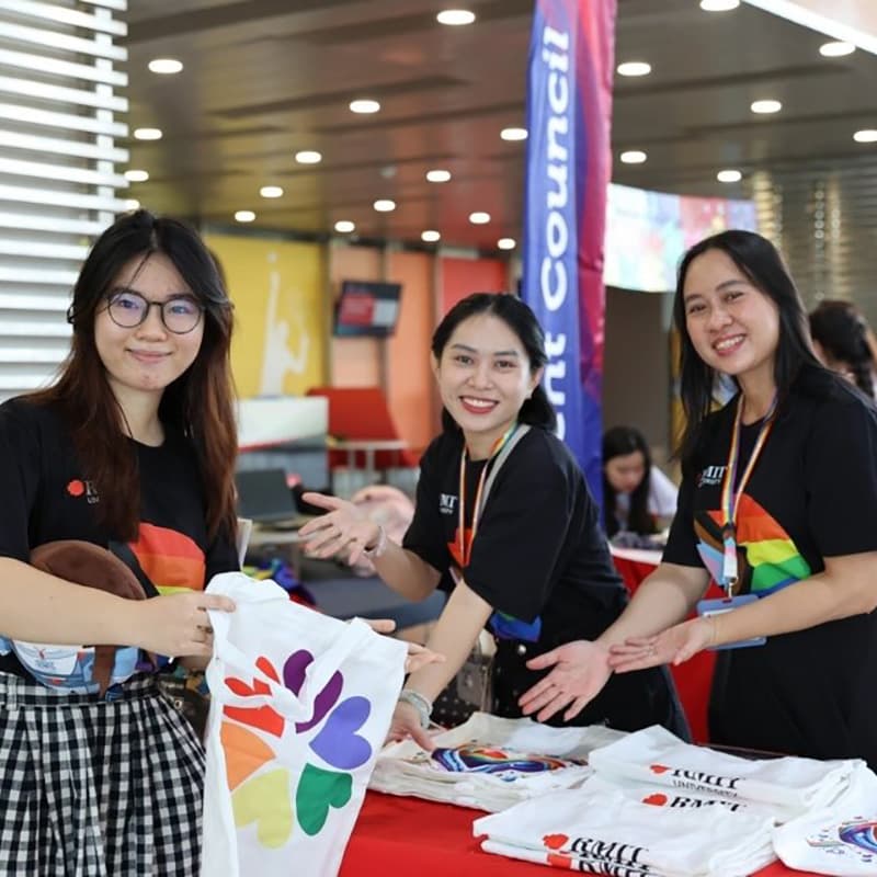 Three students at an RMIT event smiling and displaying white T-shirts with a rainbow heart design at a stall, with “Student Council” signage in the background.