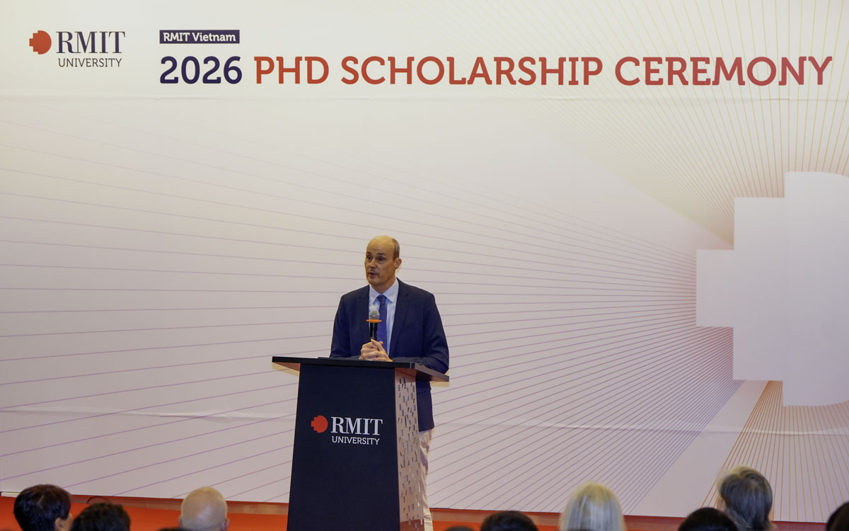 A speaker stands at an RMIT University lectern during the RMIT Vietnam 2026 PhD Scholarship Ceremony, with an event backdrop and audience seated in front.