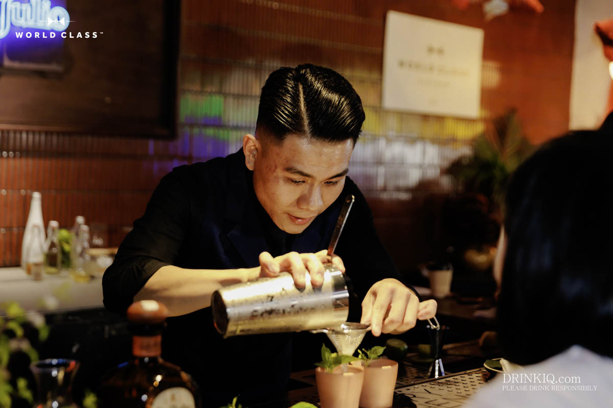 Một người đang pha chế đồ uống sau quầy ba, rót từ bình lắc kim loại vào ly.