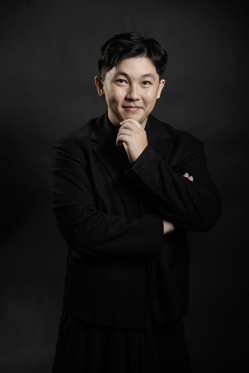 A person dressed in a dark suit poses against a plain dark background, standing with one arm crossed and the other hand raised near their chin.