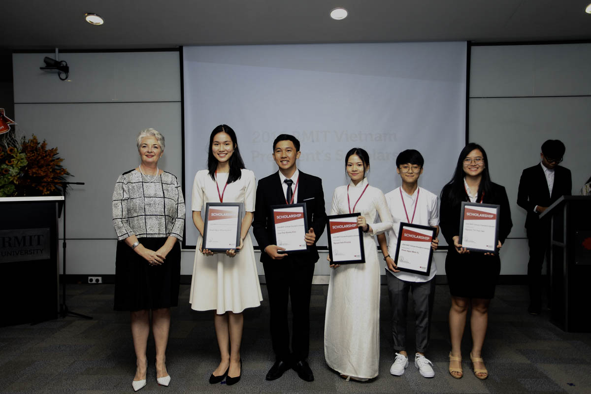 A group of six people stand in a row at an indoor award ceremony, each holding a framed certificate. A presentation screen and podiums are visible in the background.