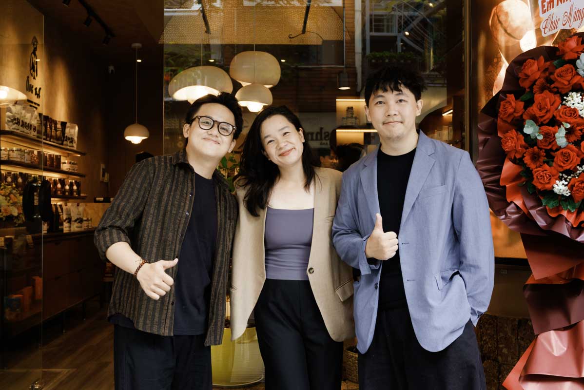 Three people stand together in front of a warmly lit storefront, with shelves, hanging lights, and café-style decor visible inside. Two individuals give a thumbs-up gesture, and a floral arrangement is displayed to the right side of the entrance.