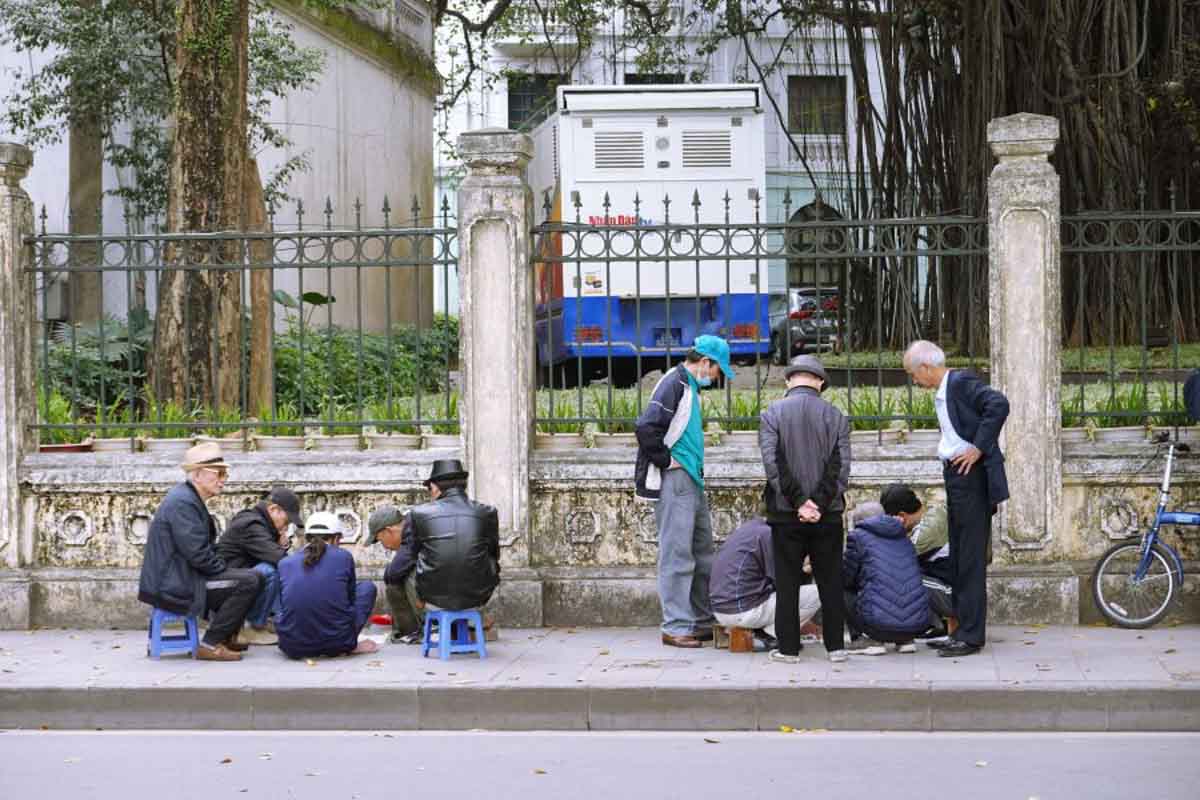 Group of older adults gathered on a sidewalk, sitting and standing around a street-side game near a fenced park. 