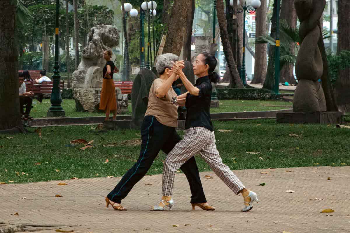 Two older women dancing on a paved path in a park surrounded by trees and sculptures.