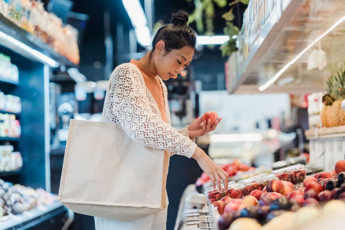 Person selecting fresh fruit in a supermarket produce section while carrying a reusable tote bag.