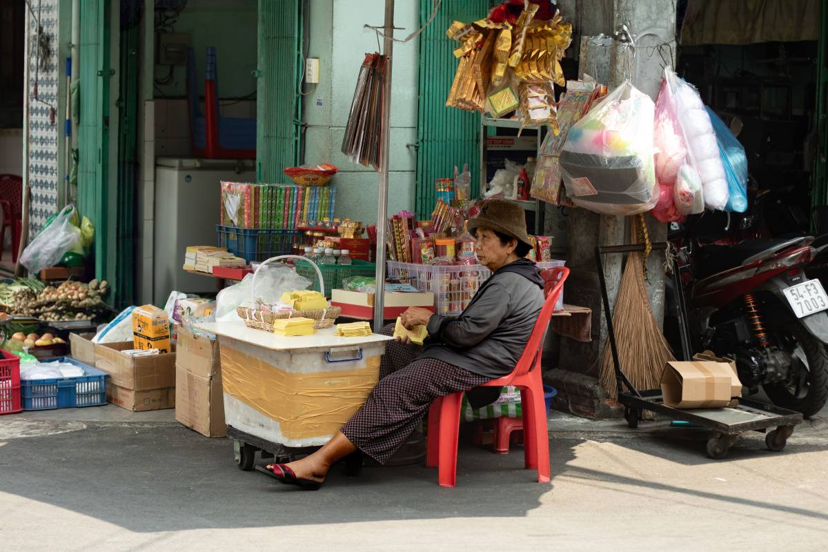 Street-side shop with a vendor seated under the shadow of an umbrella beside a display of household goods, snacks, and packaged items in a residential neighborhood.