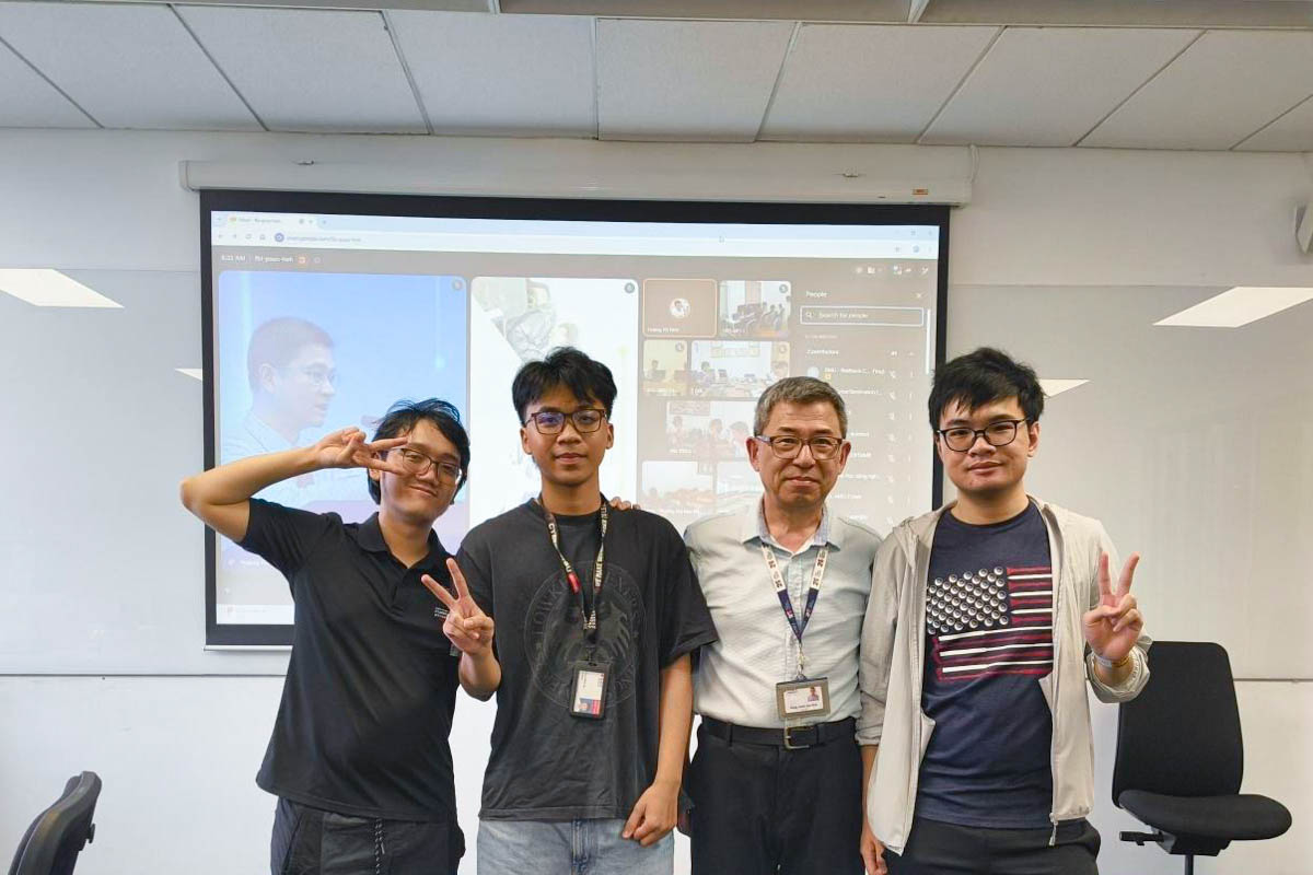 Four people posing in a classroom in front of a presentation screen displaying an online meeting.