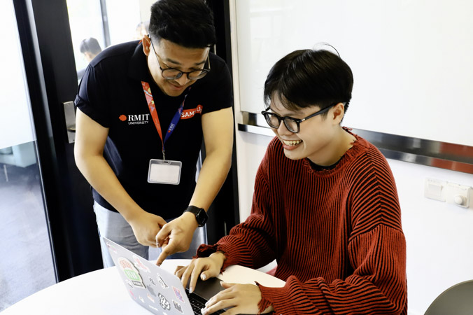 Two people at RMIT University collaborate at a table, one pointing at a laptop screen while the other sits and works, with stickers visible on the laptop in a bright indoor space.
