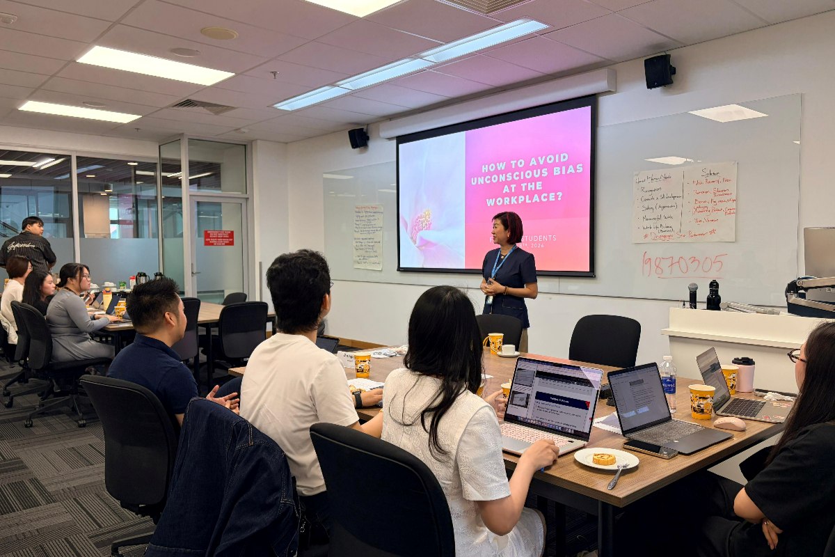 A lecturer stands at the front of an RMIT classroom delivering a presentation titled “How to avoid unconscious bias at the workplace?” while students sit at tables with laptops and notes, listening and taking part in the session.