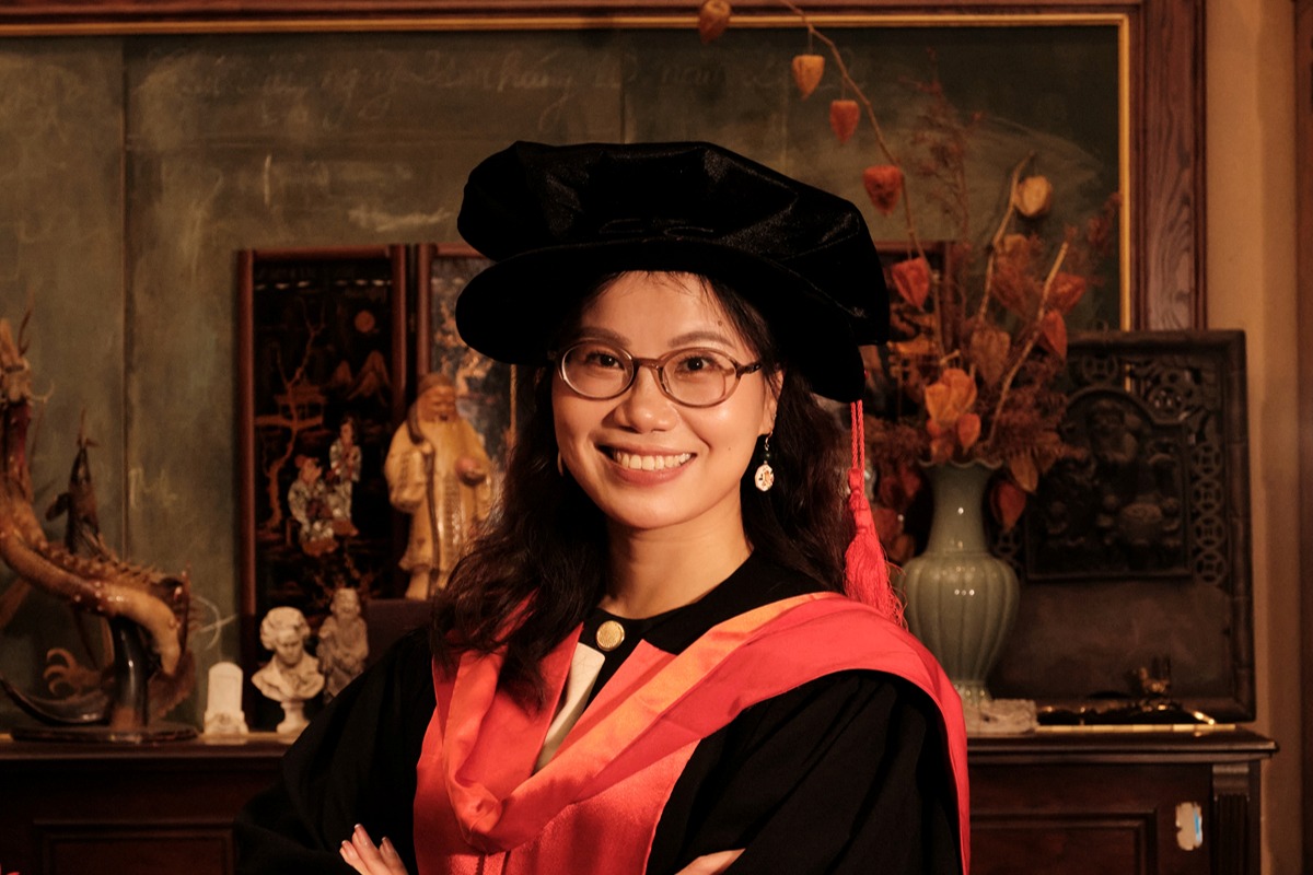 Dr Pham Thanh Hang wearing academic regalia and glasses stands indoors with crossed arms, in front of ornamental decor and art.