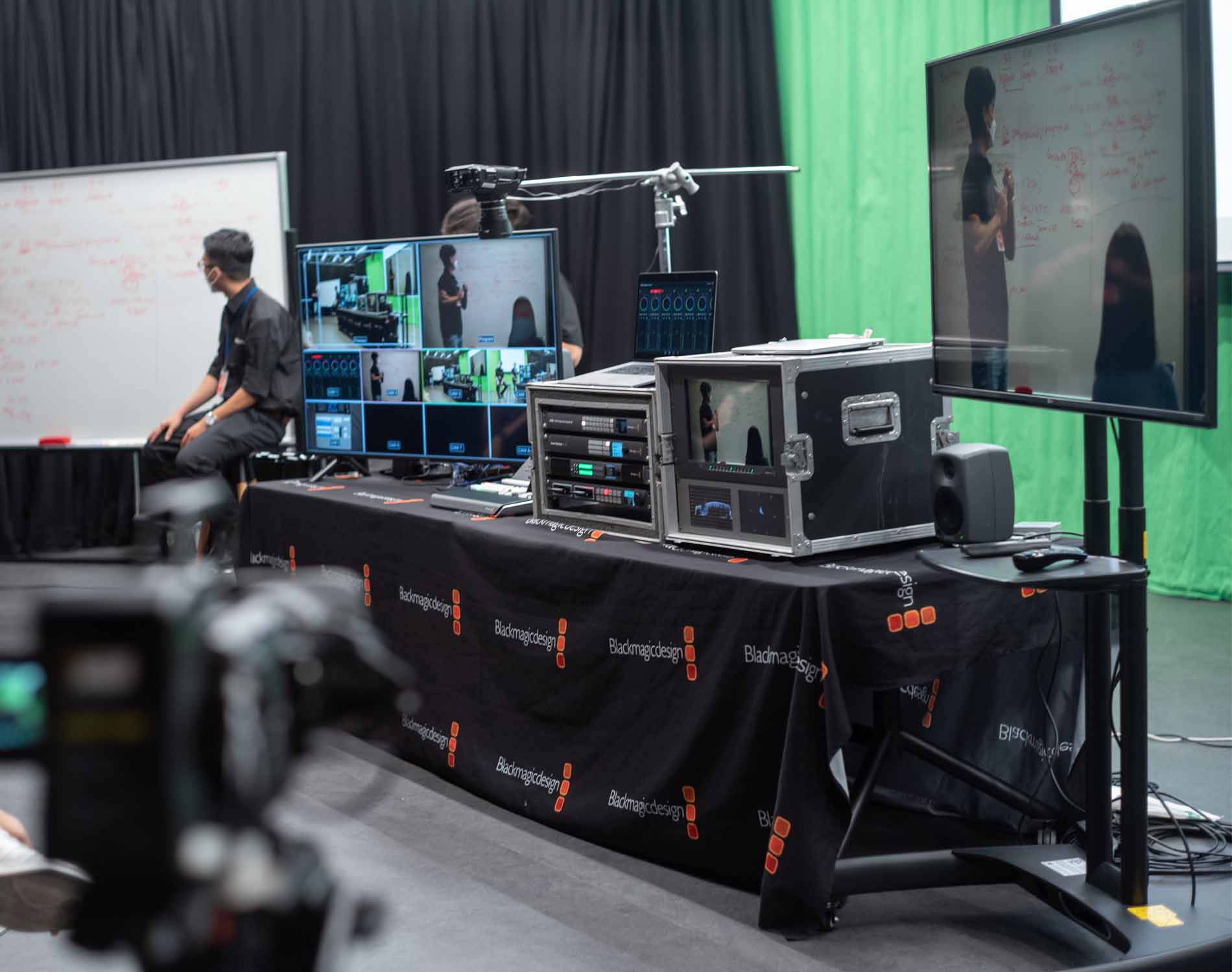 A media production setup with multiple monitors, video equipment and a technician working near a whiteboard in a studio.