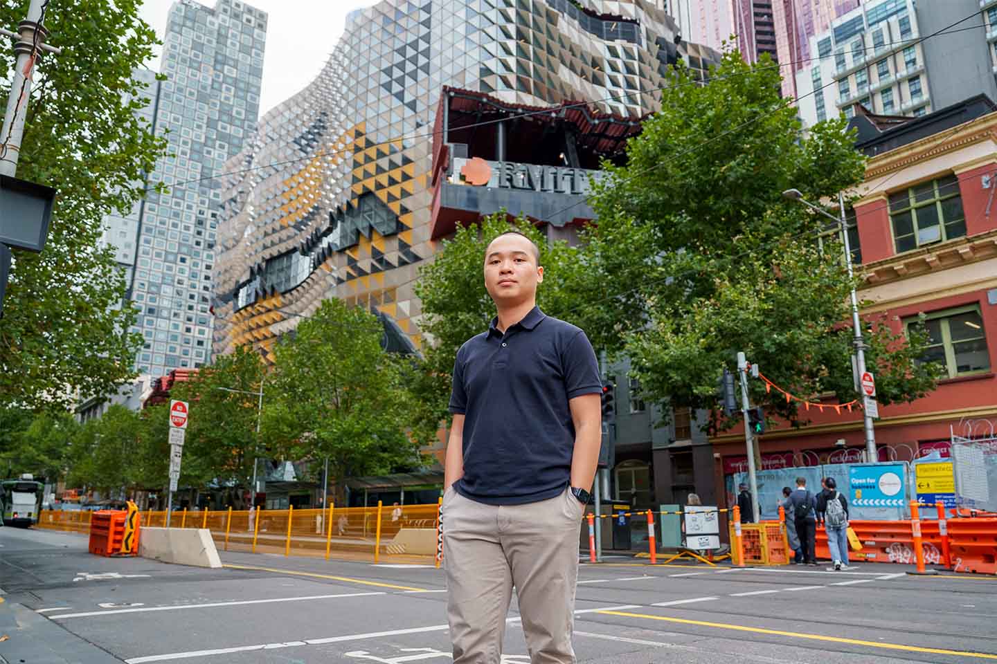 Photo of Ho Le Minh Thach, RMIT Vietnam's alumnus, standing on a city street in front of the distinctively designed RMIT University building in Melbourne, surrounded by urban greenery and construction barriers.