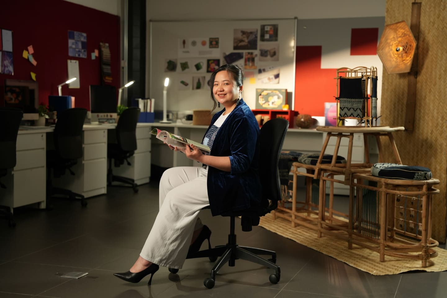Photo of Nguyen Huyen Chau, RMIT Vietnam's alumna, individual sitting in a creatively decorated office space, holding a magazine, with various design elements and materials displayed in the background.