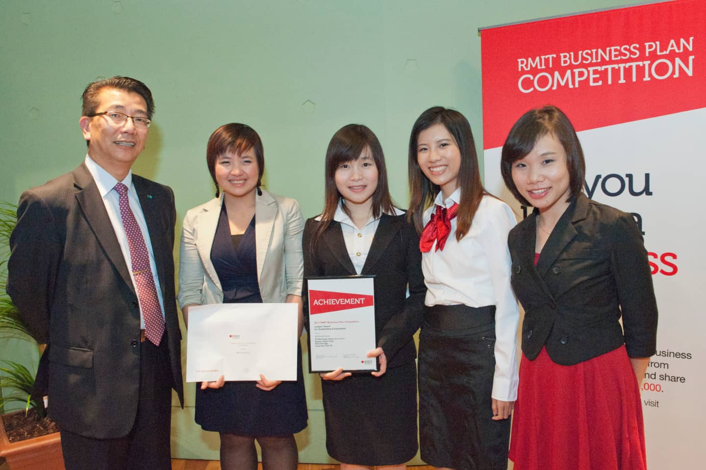 Group of participants at the RMIT Business Plan Competition, holding certificates of achievement, in front of a banner promoting business idea development.