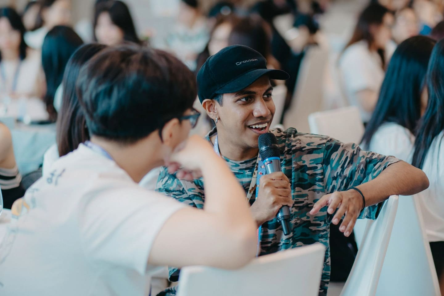 A lively event setting with attendees engaged in conversation, one holding a microphone, suggesting an interactive or discussion session.