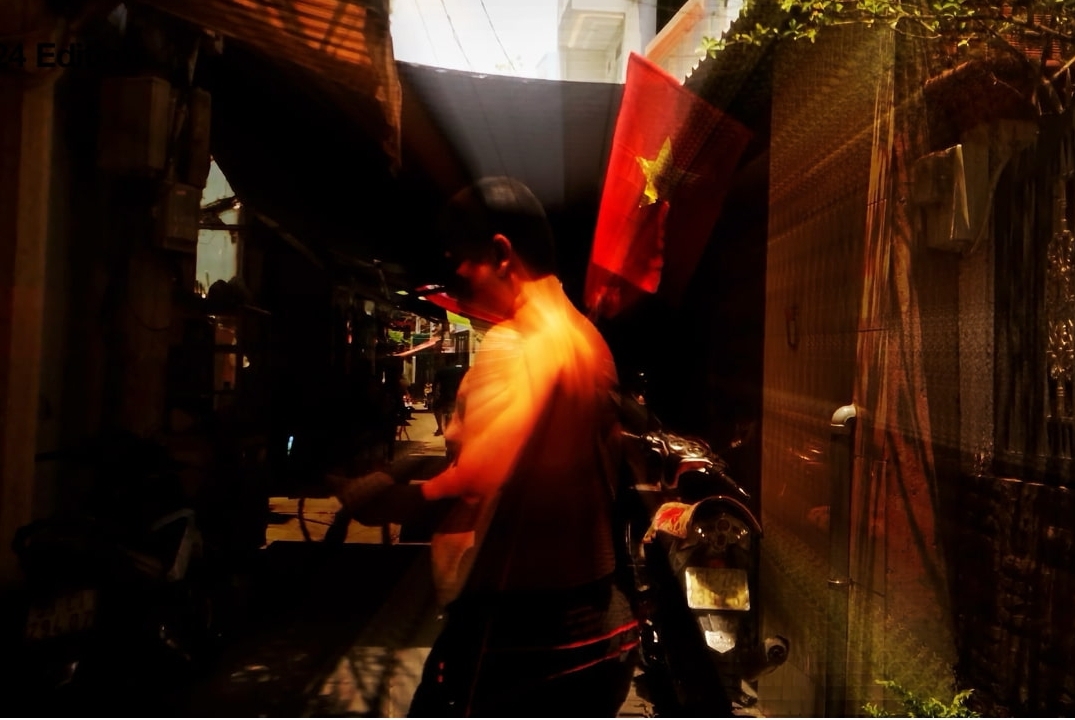 A person moves through a dimly lit alley with sunlight streaming in, a vivid Vietnamese flag with a yellow star is visible in the background, and motorbikes are parked along the sides.