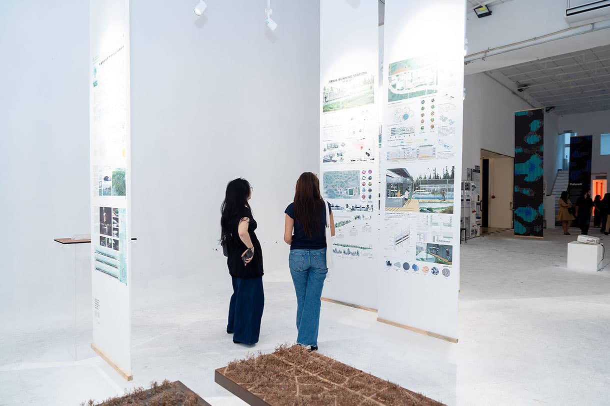 Two people are standing in a modern, white exhibition space, viewing large display boards with architectural drawings and diagrams. Brown sculptural pieces are visible on the floor.