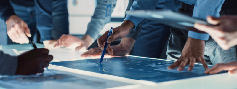 Stock photo of a team gathering around a table with a glowing digital display reviewing a technical blueprint or research data. Hands are pointing and interacting with the screen