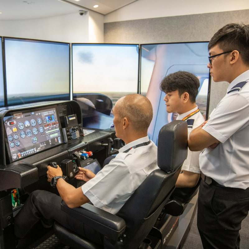 Photo of a group of students and a lecturer using a flight simulator with multiple screens displaying cockpit controls and an aerial view.