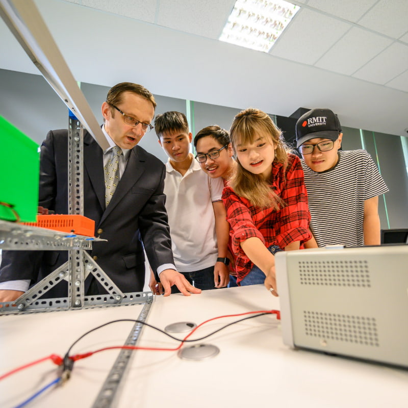 Photo of a group of students and a lecturer engaged in a collaborative engineering or technology project around a table with electronic equipment and structural components.