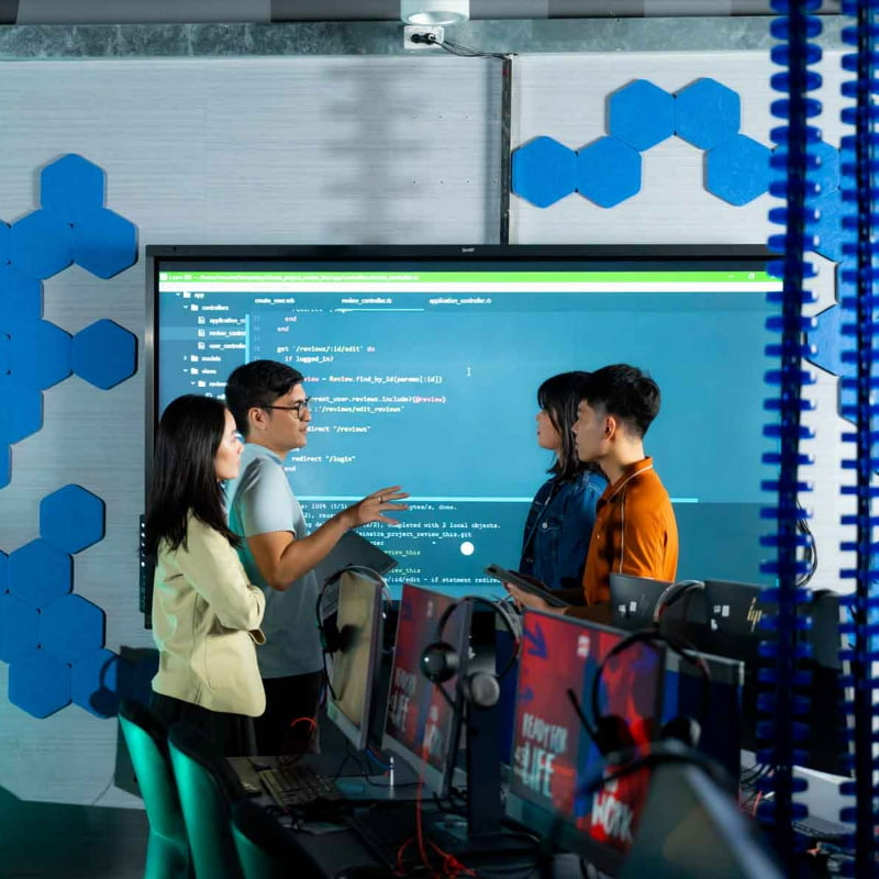 Photo of a group of people engaged in a discussion in a tech-savvy room with a large screen displaying code in the background, reflecting collaboration in a programming or software development environment.