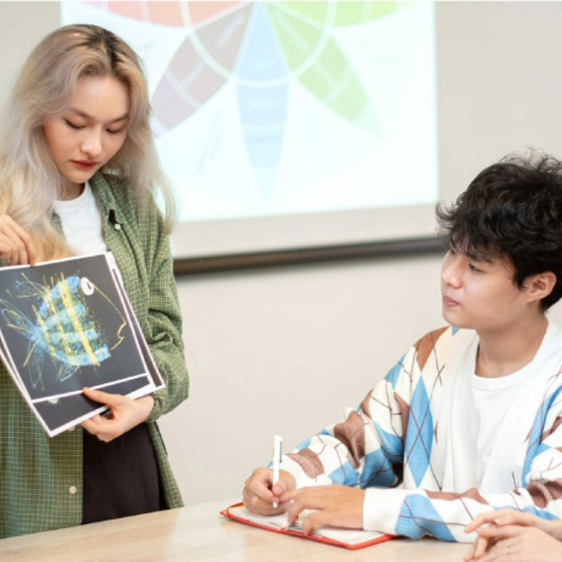 Photo of a student presenting a creative artwork or design to another student who is taking notes, in a meeting or brainstorming session setting with a color wheel on a screen in the background.
