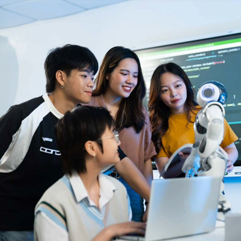 Photo of a group of four students gathered around a desk with a laptop and humanoid robot in a tech-focused environment, suggesting teamwork on a project involving programming or robotics.