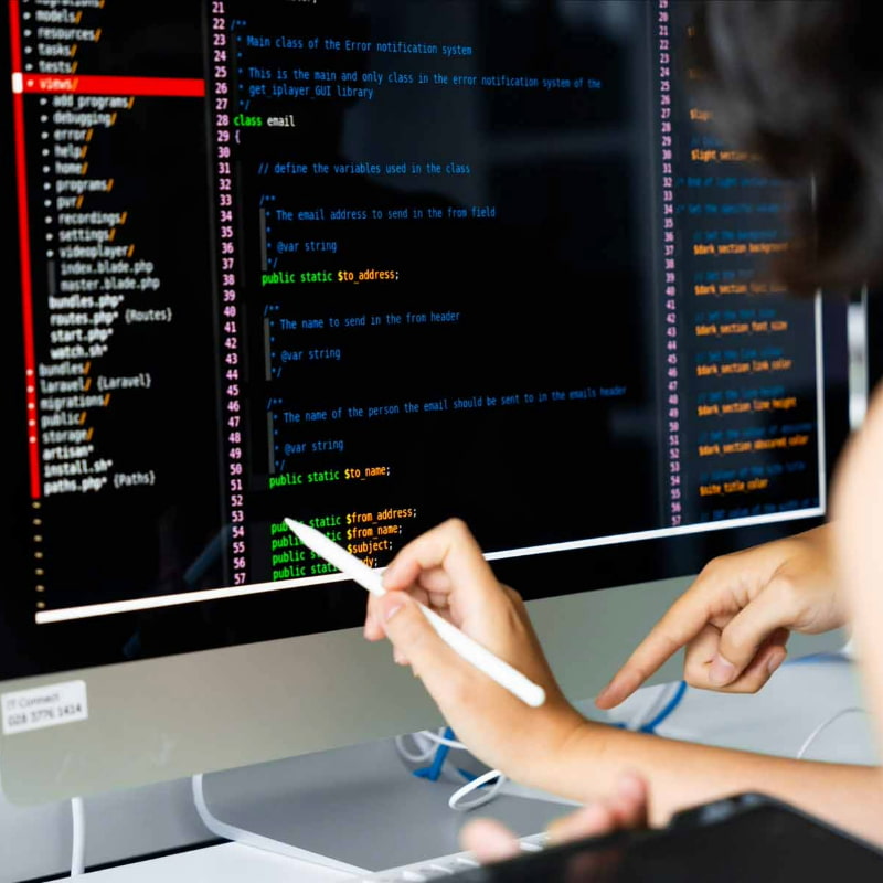 Photo of two people pointing at a large computer screen displaying colorful lines of code, indicating collaboration or discussion on programming tasks.