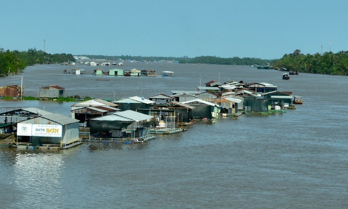 Group of floating houses and structures sit on a wide, calm river.