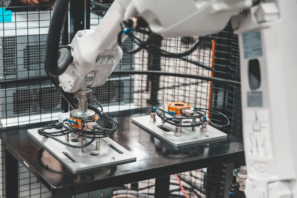 Photo of a robotic arm operating in an industrial setting, working on two metal plates with complex wiring on a table, 
