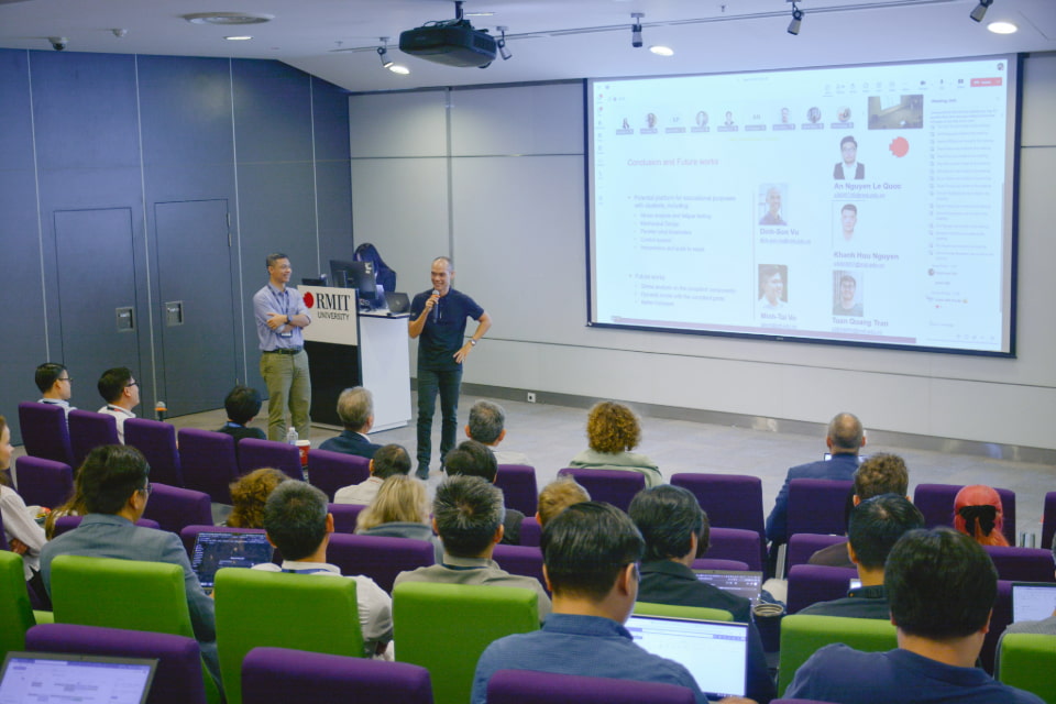 A snapshot of an event where two presenters standing in front of an audience in a lecture hall. A large screen behind them displays a presentation with profile photos and text. The audience is seated in rows with purple and green chairs, some using laptops.