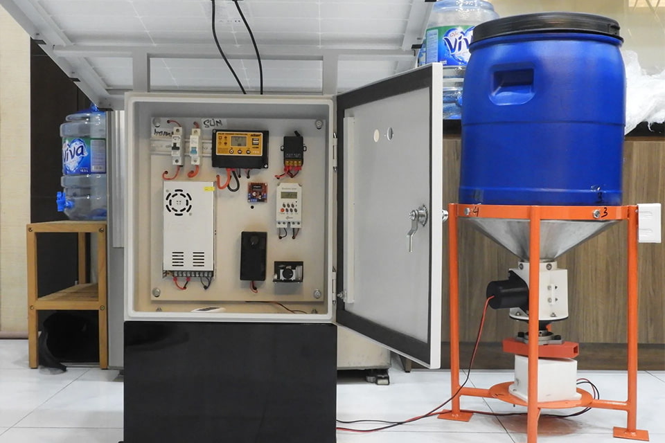 Real life prototype of a smart shrimp feeding system setup featuring a control panel with electronic components inside an open cabinet, connected to a blue feed hopper with a red stand