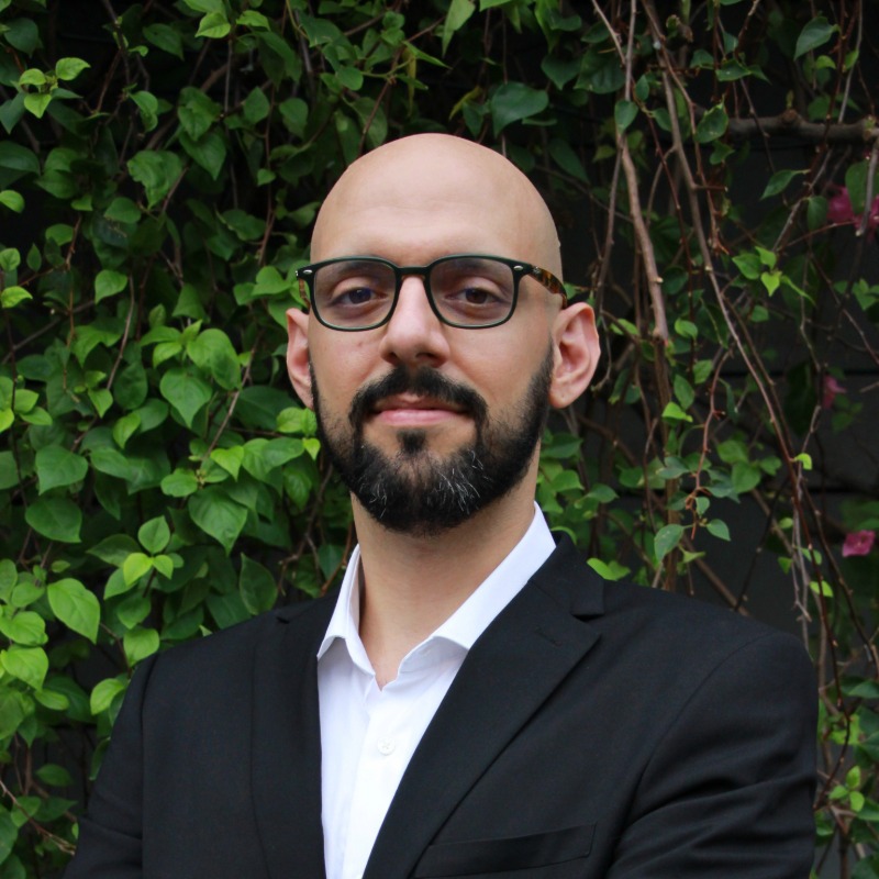 Man with glasses and a beard, wearing a dark suit and white shirt, stands with arms crossed in front of a leafy green background.