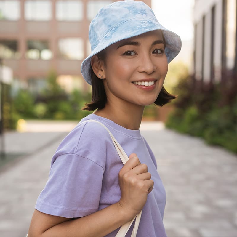 A young woman smiling outdoors, wearing a light blue bucket hat and lavender T-shirt, carrying a canvas bag over her shoulder, with a blurred urban background including buildings and greenery.