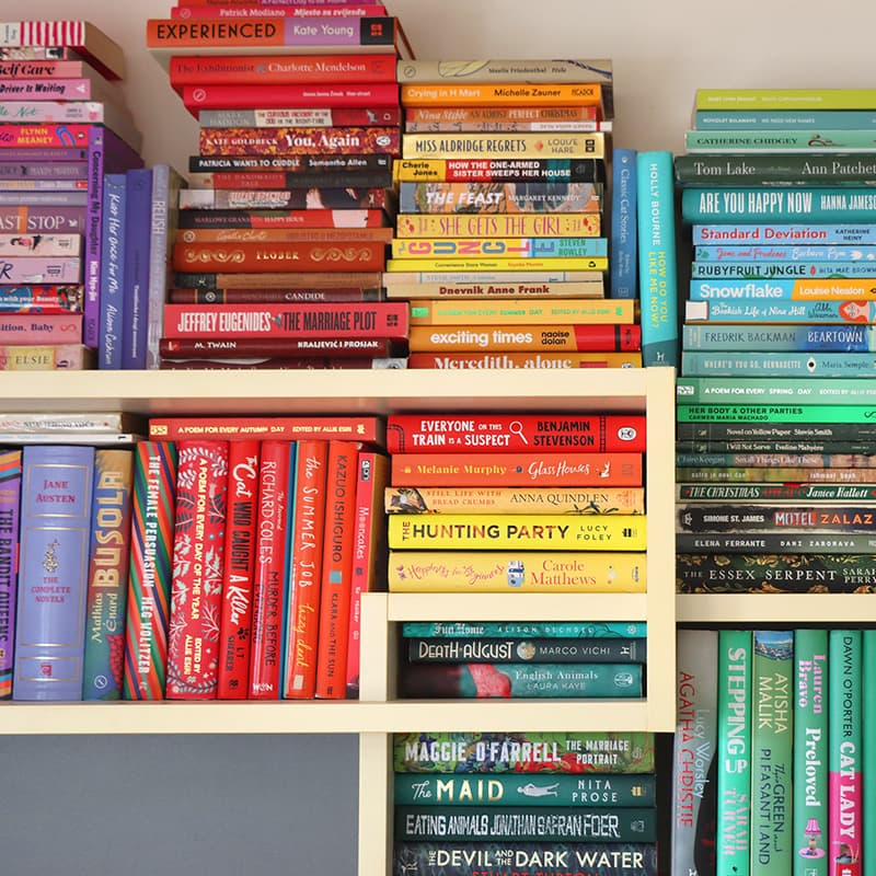 Colourful shelves filled with books arranged by spine colour, creating a rainbow effect with sections of red, orange, yellow, green, blue and purple book covers. The book titles and authors are visible.