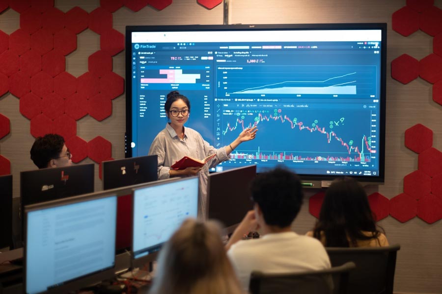 Photo of a female student presenting a stock market graph with fluctuating lines on a large screen to other students in a computer laboratory at RMIT Vietnam