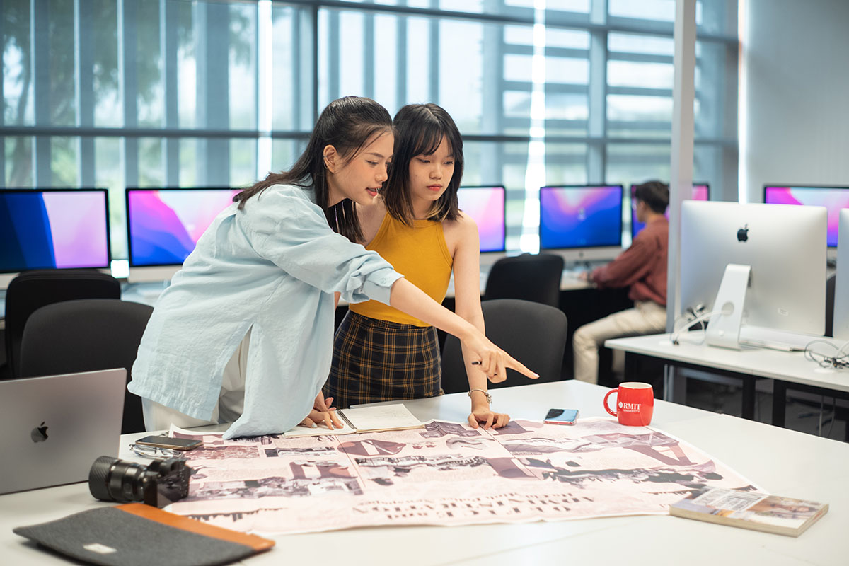Hai sinh viên trong phòng máy tính xem xét bản thiết kế in khổ lớn trên bàn, bên cạnh có máy ảnh và laptop