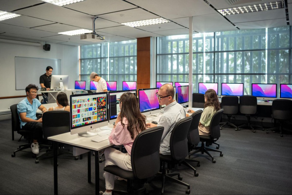 Sinh viên trong phòng máy tính làm dự án thiết kế đồ họa, ngồi tại bàn với màn hình lớn hiển thị bố cục màu sắc