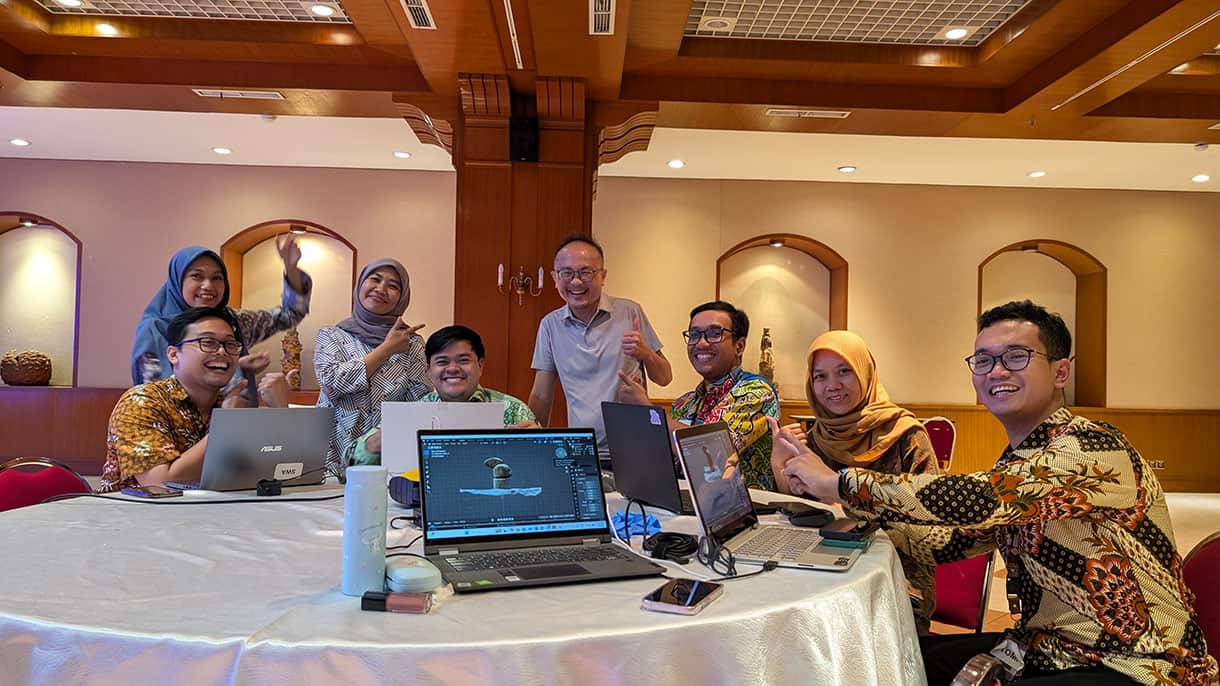 A group of people sitting and standing around a round table with laptops, smiling and posing cheerfully in a warmly-lit meeting room.
