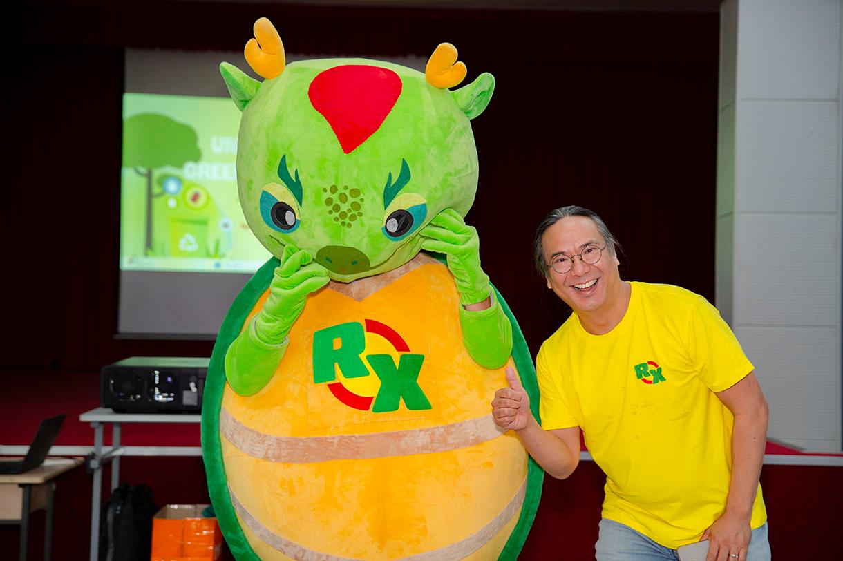 Lecturer Nhan Nguyen in a yellow RX t-shirt stands next to a large green and yellow mascot costume with dragon features, giving a thumbs up at an indoor event.